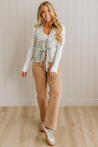 Woman wearing a floral vest, white long-sleeve shirt, tan pants, and silver shoes against a beige wall.