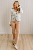 Woman wearing a floral vest over a white shirt with beige pants against a light wooden paneled wall.