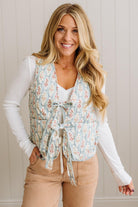 Woman wearing a floral vest over a white long-sleeve shirt and beige pants against a neutral background