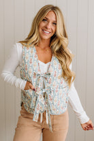 Woman wearing a floral vest over a white long-sleeve shirt and tan pants against a light wooden panel background.
