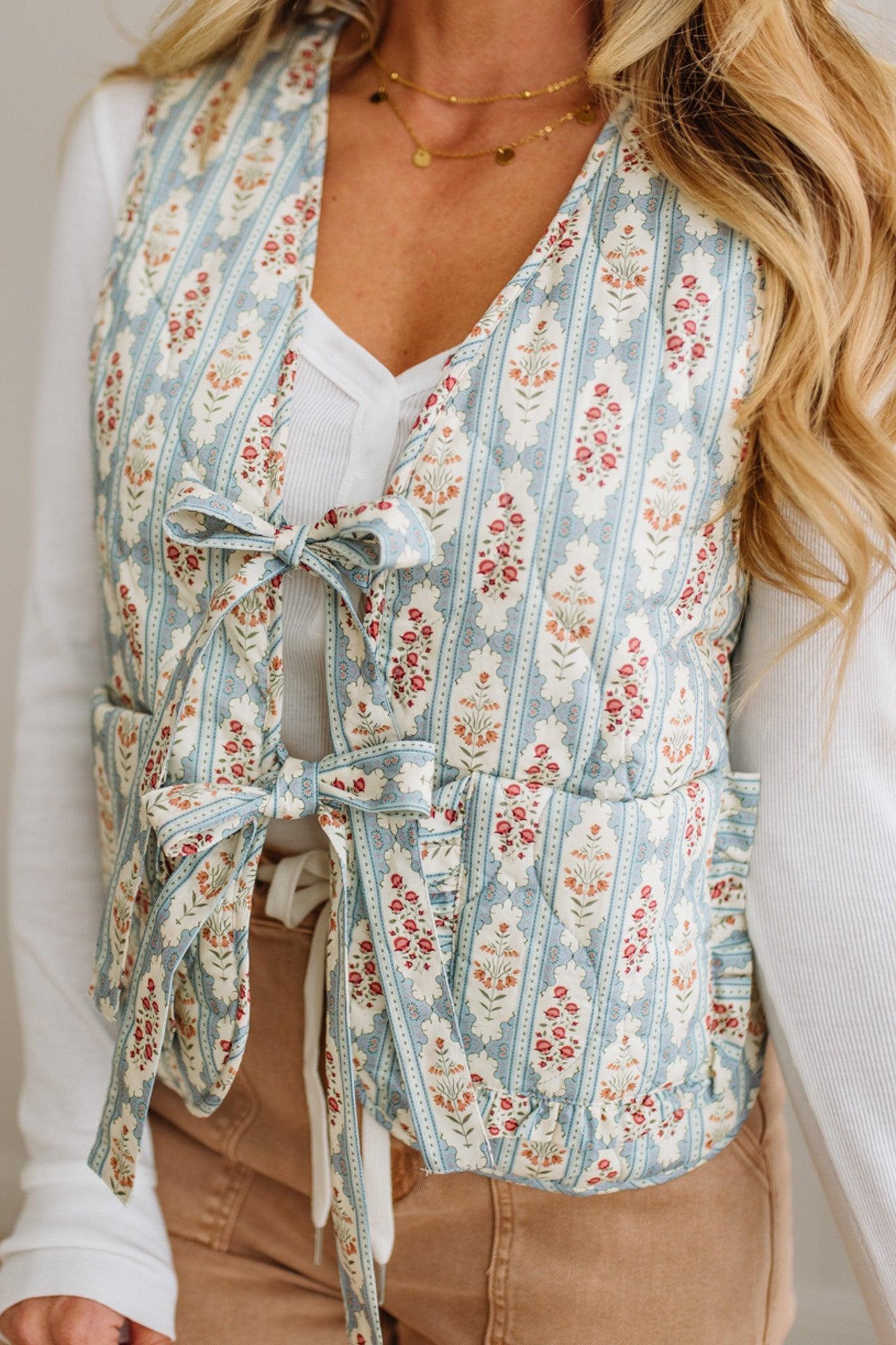 Woman wearing a floral patterned vest over a white shirt and beige pants.