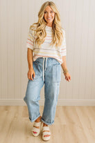 Woman wearing a striped shirt and jeans standing against a light wooden paneled wall.