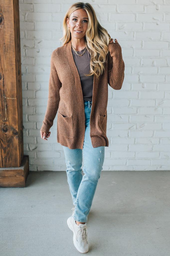 Woman wearing a brown cardigan, gray top, light blue jeans, and white sneakers in a room with a wooden cabinet and white brick wall.