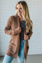 Woman wearing a brown cardigan over a gray shirt and blue jeans against a white brick wall.