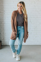 Woman wearing a brown cardigan, gray shirt, blue jeans, and white sneakers against a white brick wall.