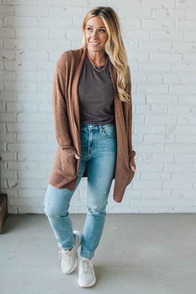 Woman wearing a brown cardigan, gray shirt, blue jeans, and white sneakers against a white brick wall.