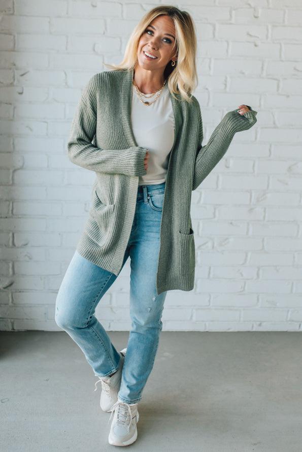 Woman wearing a green cardigan, white shirt, blue jeans, and white sneakers against a white brick wall.