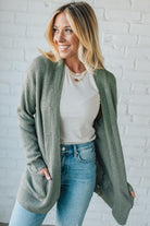 Woman wearing a green cardigan over a white shirt and blue jeans against a white brick wall.