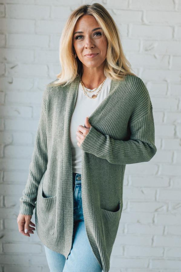 Woman wearing a green cardigan over a white shirt with light blue jeans against a white brick wall.