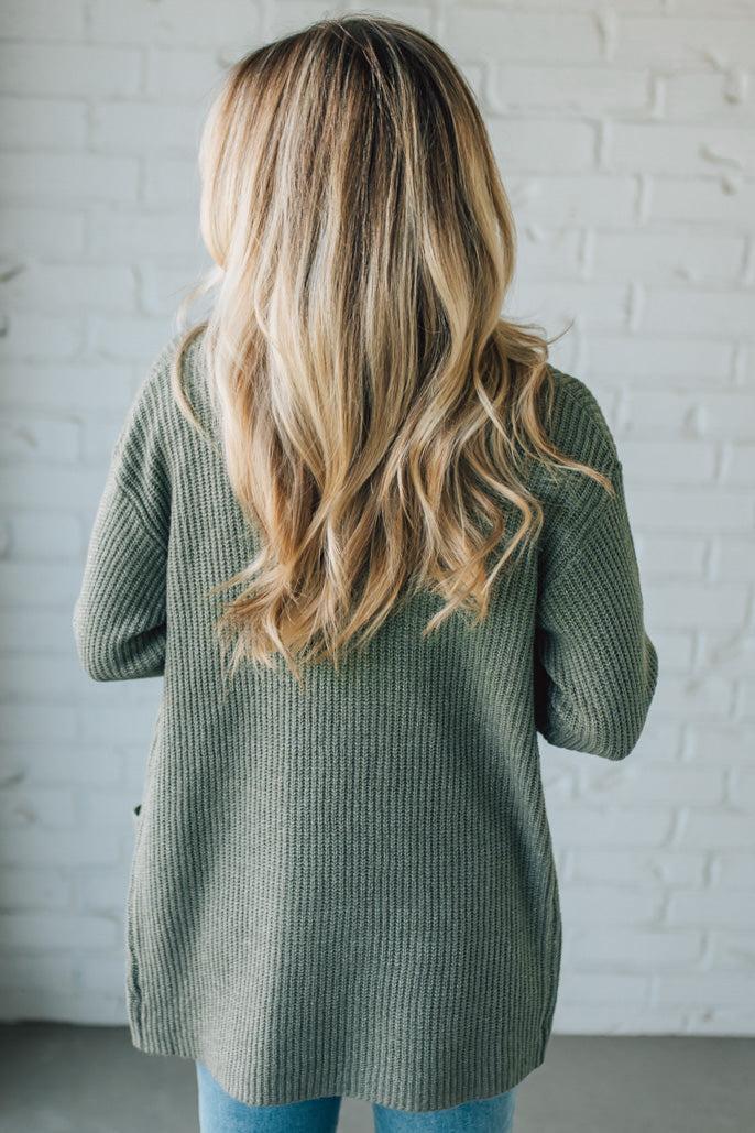 Person wearing a green knitted cardigan against a white brick wall.