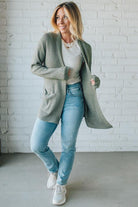 Woman wearing a green cardigan, white shirt, blue jeans, and white sneakers against a white brick wall.