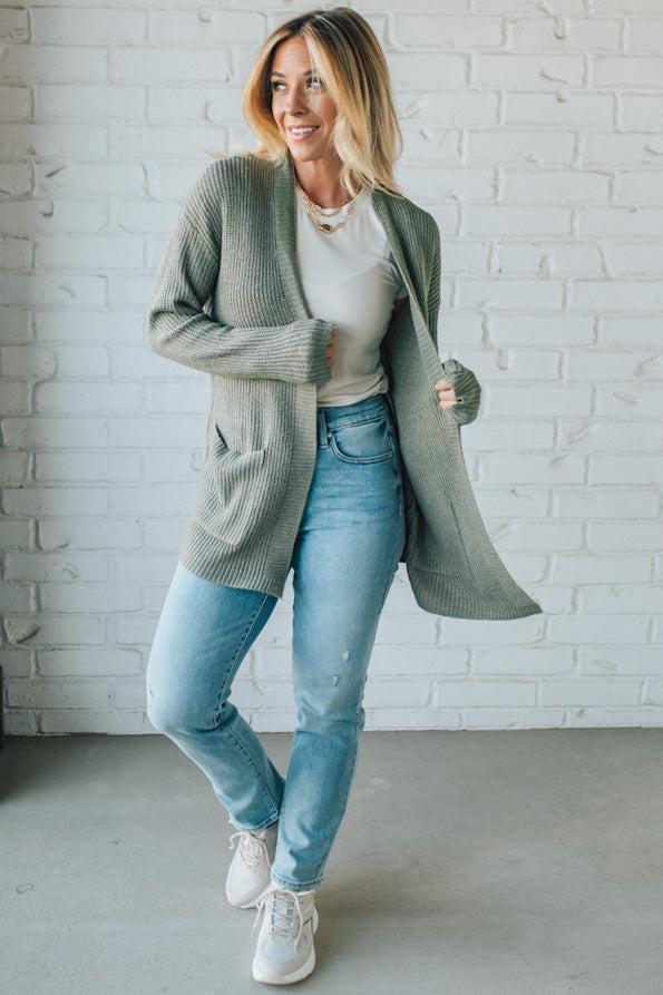 Woman wearing a green cardigan, white shirt, blue jeans, and white sneakers against a white brick wall.