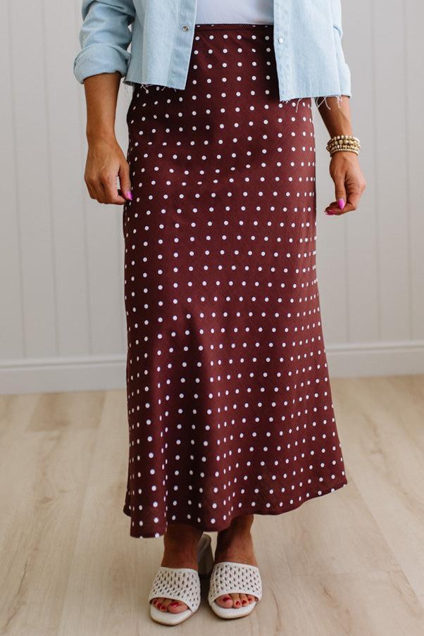Person wearing a maroon polka dot skirt with a light blue denim jacket and white sandals on a wooden floor.