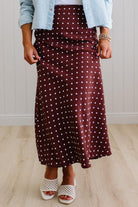 Person wearing a burgundy polka dot skirt with a light blue jacket and white sandals on a wooden floor.