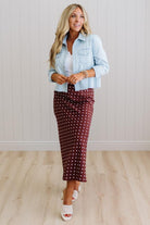 Woman wearing a light blue denim jacket over a white shirt and burgundy polka dot skirt on a plain background