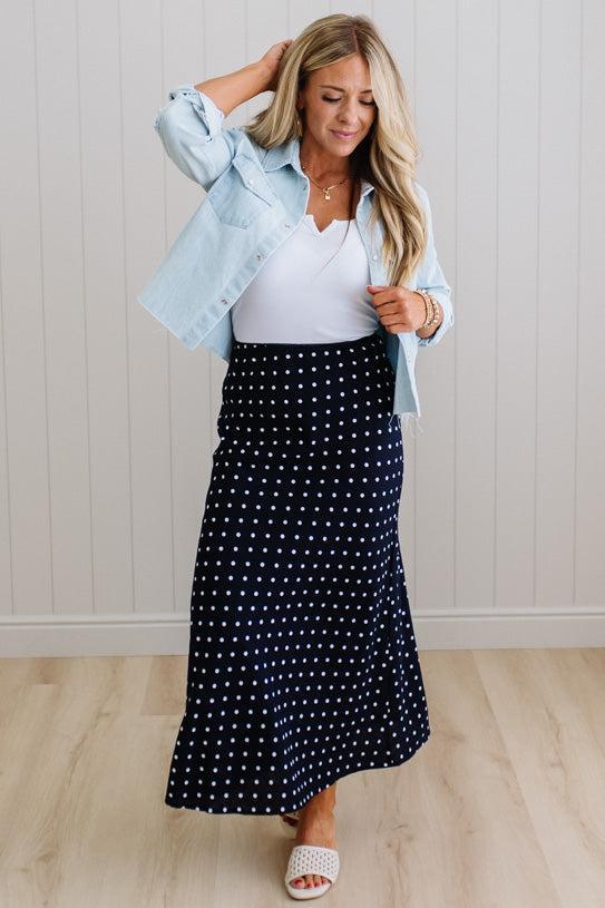 Woman wearing a navy polka dot skirt with a white top and light blue jacket indoors.