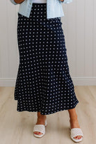 Person wearing a black polka dot skirt with a light blue shirt and white sandals on a wooden floor.