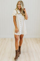 A woman modeling a short-sleeve cream button-front dress with small brown polka dots, paired with brown ankle boots, standing in a room with light wood floors and white paneled walls.