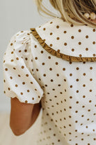 Close-up of a child wearing a cream-colored dress with brown polka dots, featuring a ruffled collar and puff sleeves.