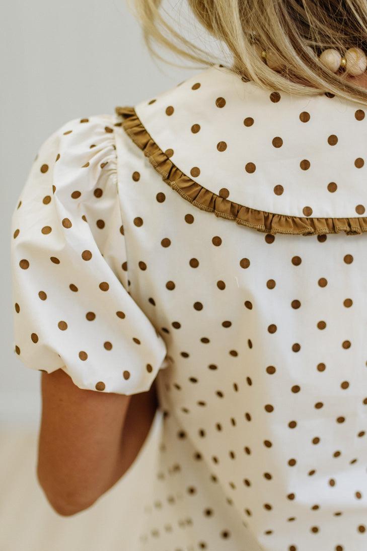 Close-up of a child wearing a cream-colored dress with brown polka dots, featuring a ruffled collar and puff sleeves.