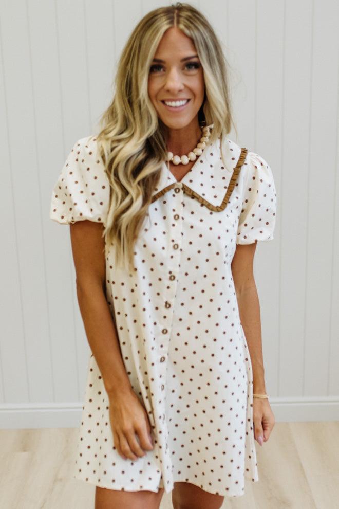 A woman wearing a short-sleeve cream dress with brown polka dots and a brown decorative collar, standing against a white paneled wall.