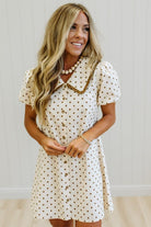 A woman wearing a cream-colored short-sleeve dress with brown polka dots and a brown collar trim, standing against a white paneled wall.