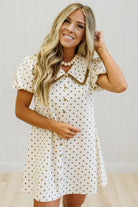 A woman modeling a short-sleeve cream dress with brown polka dots, a brown collar, and brown buttons, standing against a light-colored paneled wall.