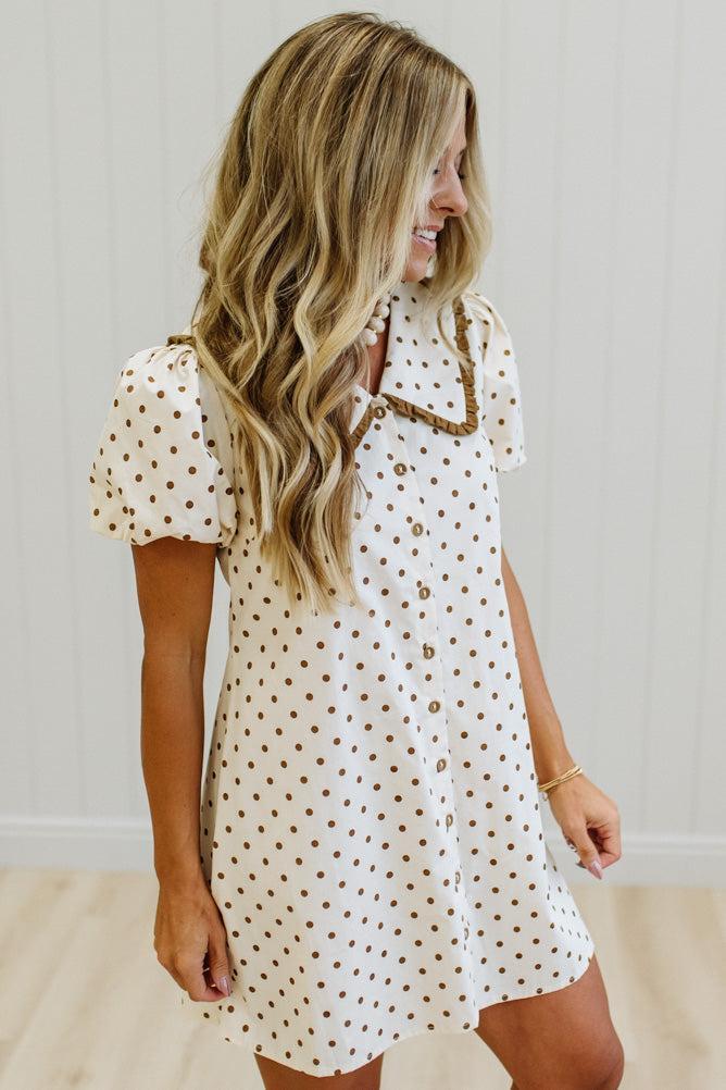A woman wearing a short-sleeve white button-up dress with brown polka dots and a brown collar trim, standing against a white paneled wall.