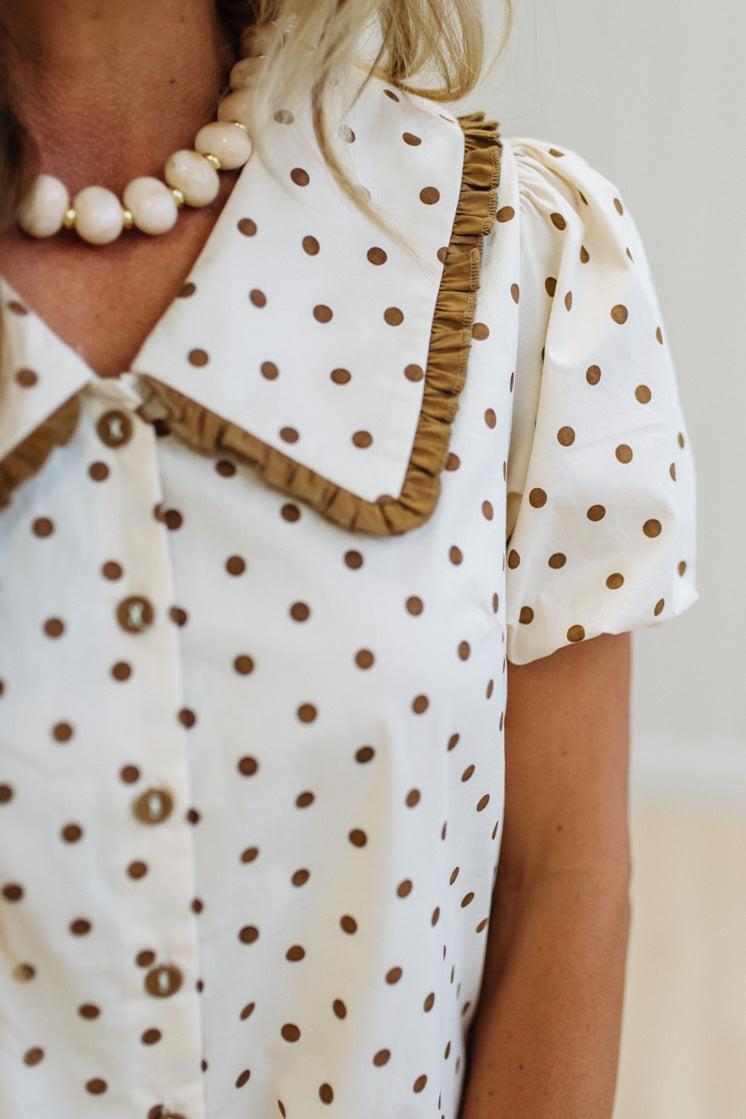 Close-up of a person wearing a white blouse with brown polka dots and a ruffled collar, paired with a pearl necklace.