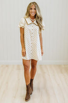 A woman modeling a short-sleeve white button-front dress with a small tan polka dot pattern, a tan collar, and brown ankle boots, standing in a room with light wood floors and white paneled walls.