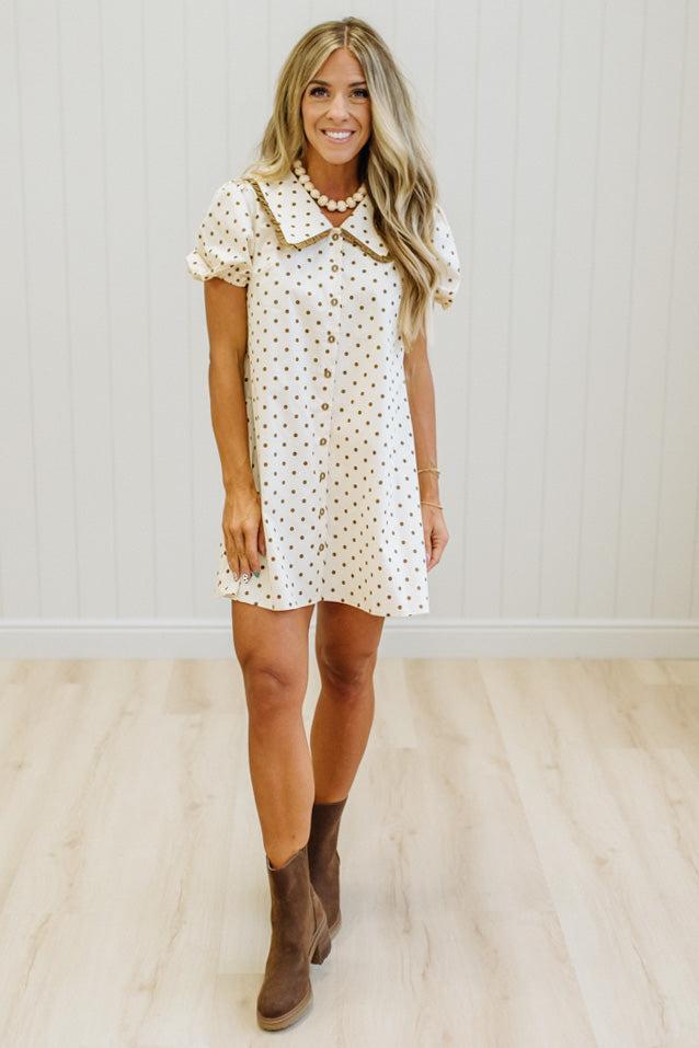 A woman modeling a short-sleeve white button-front dress with a small tan polka dot pattern, a tan collar, and brown ankle boots, standing in a room with light wood floors and white paneled walls.
