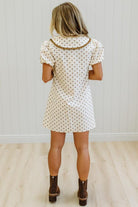 A woman modeling a short-sleeve cream dress with brown polka dots, a brown collar, and brown buttons, standing against a light-colored paneled wall.