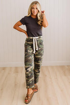 A woman modeling camouflage drawstring pants, a dark charcoal t-shirt, and brown slide sandals in a room with light wood floors and white paneled walls.