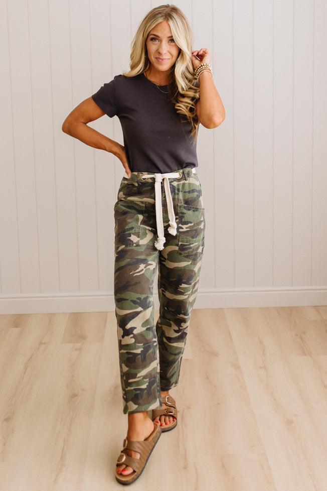 A woman modeling camouflage drawstring pants, a dark charcoal t-shirt, and brown slide sandals in a room with light wood floors and white paneled walls.