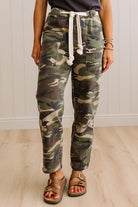 A person wearing camouflage jogger pants with a white drawstring, paired with brown sandals, standing against a white paneled wall and light wood floor.