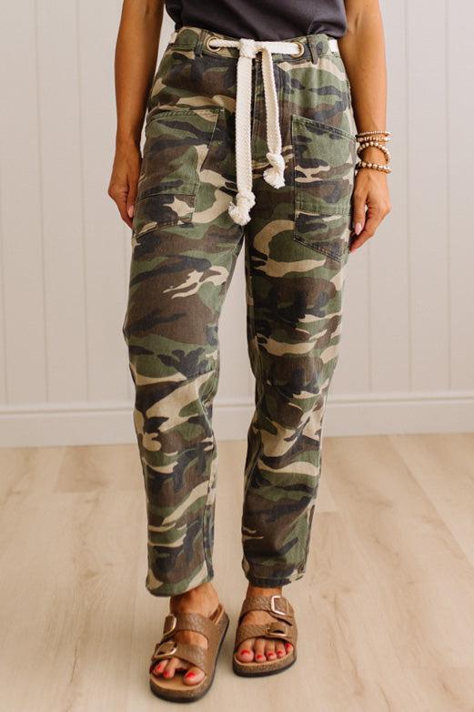 A person wearing camouflage jogger pants with a white drawstring, paired with brown sandals, standing against a white paneled wall and light wood floor.