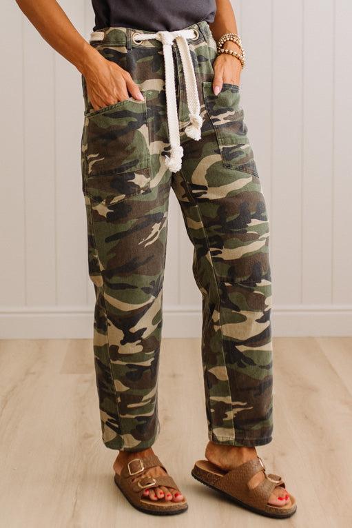 A person wearing camouflage drawstring pants with side pockets, paired with brown double-strap sandals, standing against a white paneled wall and light wood floor.