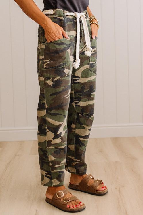 A person wearing camouflage cargo-style pants with a white drawstring, standing in a room with light wood flooring and white paneled walls.