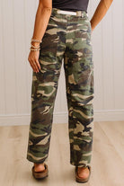 A person wearing camouflage pants with a white belt, paired with brown wedge sandals and bracelets, standing in a room with light wood floors and white paneled walls.