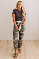 A woman modeling camouflage cargo-style pants with a white drawstring, paired with a dark gray t-shirt and brown sandals, standing against a white paneled wall and light wood floor.