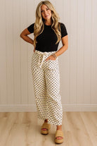 Woman wearing a black top and white polka dot pants standing against a light wooden paneled wall.