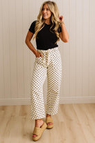 Woman wearing a black top and white polka dot pants standing against a light wooden paneled wall.