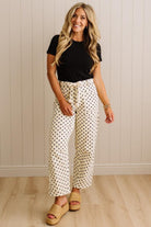Woman wearing a black top and polka dot pants standing against a light wooden paneled wall.