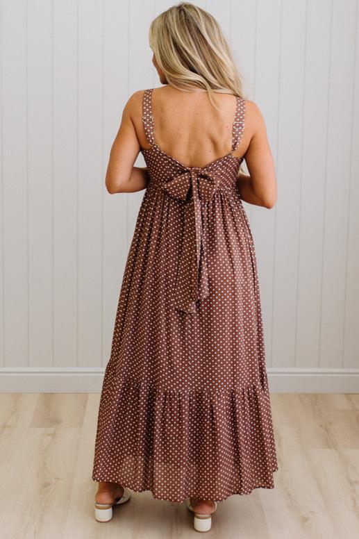 Woman wearing a brown polka dot dress with a bow detail, standing against a white wall.