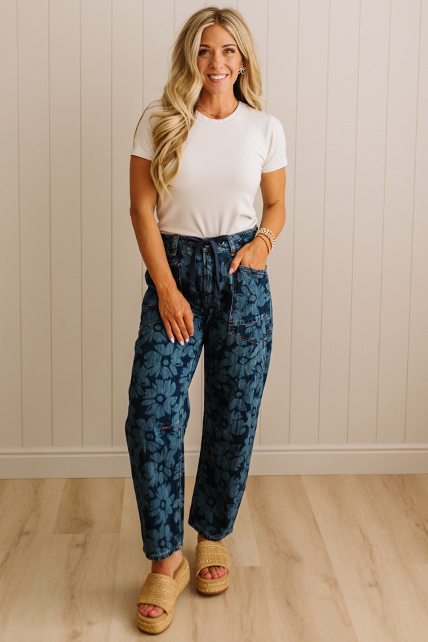 Woman wearing a white t-shirt and blue patterned pants standing against a beige wall.