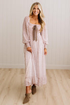 A woman modeling a light pink long-sleeve maxi dress with a tie-neck detail, standing in front of a white paneled wall and light wood floor.