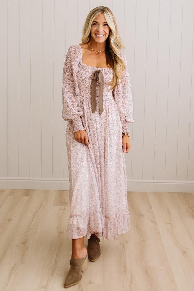 A woman modeling a light pink long-sleeve maxi dress with a tie-neck detail, standing in front of a white paneled wall and light wood floor.