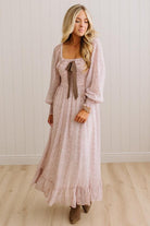 A woman wearing a long-sleeved, dusty pink textured maxi dress with a square neckline, a brown ribbon tie at the waist, and a ruffled hem, standing in front of a white paneled wall and light wood floor.