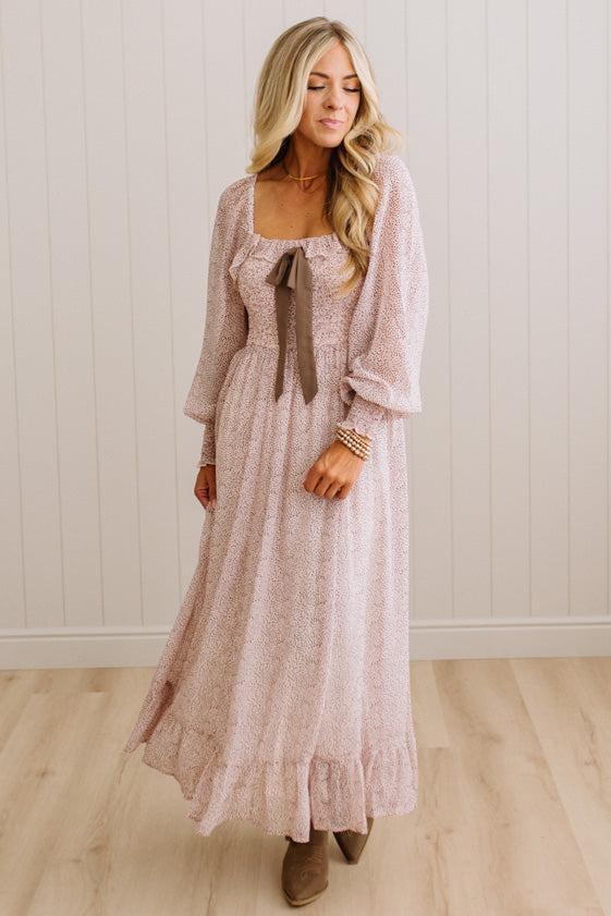 A woman wearing a long-sleeved, dusty pink textured maxi dress with a square neckline, a brown ribbon tie at the waist, and a ruffled hem, standing in front of a white paneled wall and light wood floor.