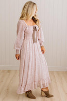 A woman modeling a long-sleeve dusty pink maxi dress with a tie-neck detail, standing in front of a white paneled wall and light wood floor.